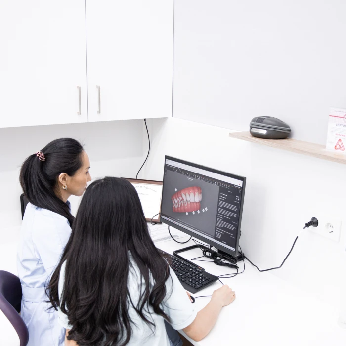 Two female dental professionals sitting at a white office desk in a clinic. They are looking at a computer monitor that shows a detailed 3D digital model of human teeth and braces.