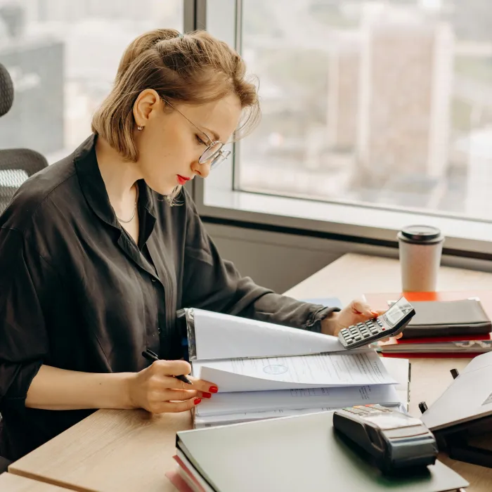 A female accountant with glasses sitting at an office desk. She is checking paper documents while holding a calculator in a modern high-rise office.