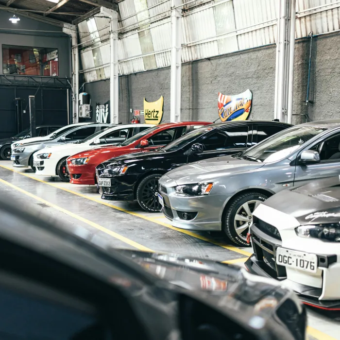A long row of modern sports cars and saloons parked neatly inside a spacious commercial warehouse garage. The vehicles, in various colours like silver, black, and red, sit under an industrial corrugated roof with bright skylights.