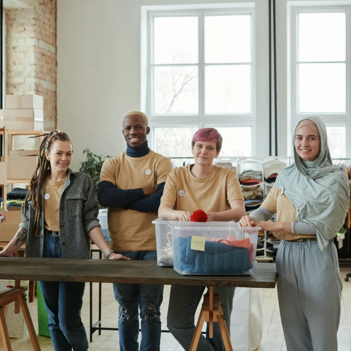 Four diverse charity volunteers standing together smiling behind a wooden table. They are wearing matching t-shirts and sorting through clothing donation boxes inside a bright community center.
