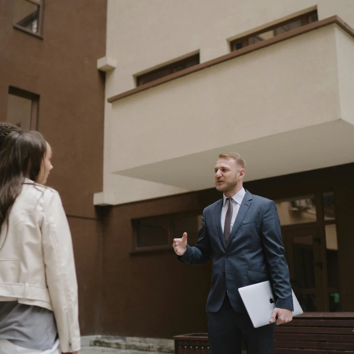 A professional estate agent in a navy blue suit talking to a client outside a modern apartment building. He is gesturing with his hand while holding a silver laptop.