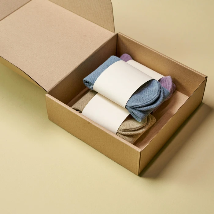 An open brown cardboard box containing neatly packed, folded pairs of socks in pastel blue, beige, and purple, each wrapped in a blank white paper label band.